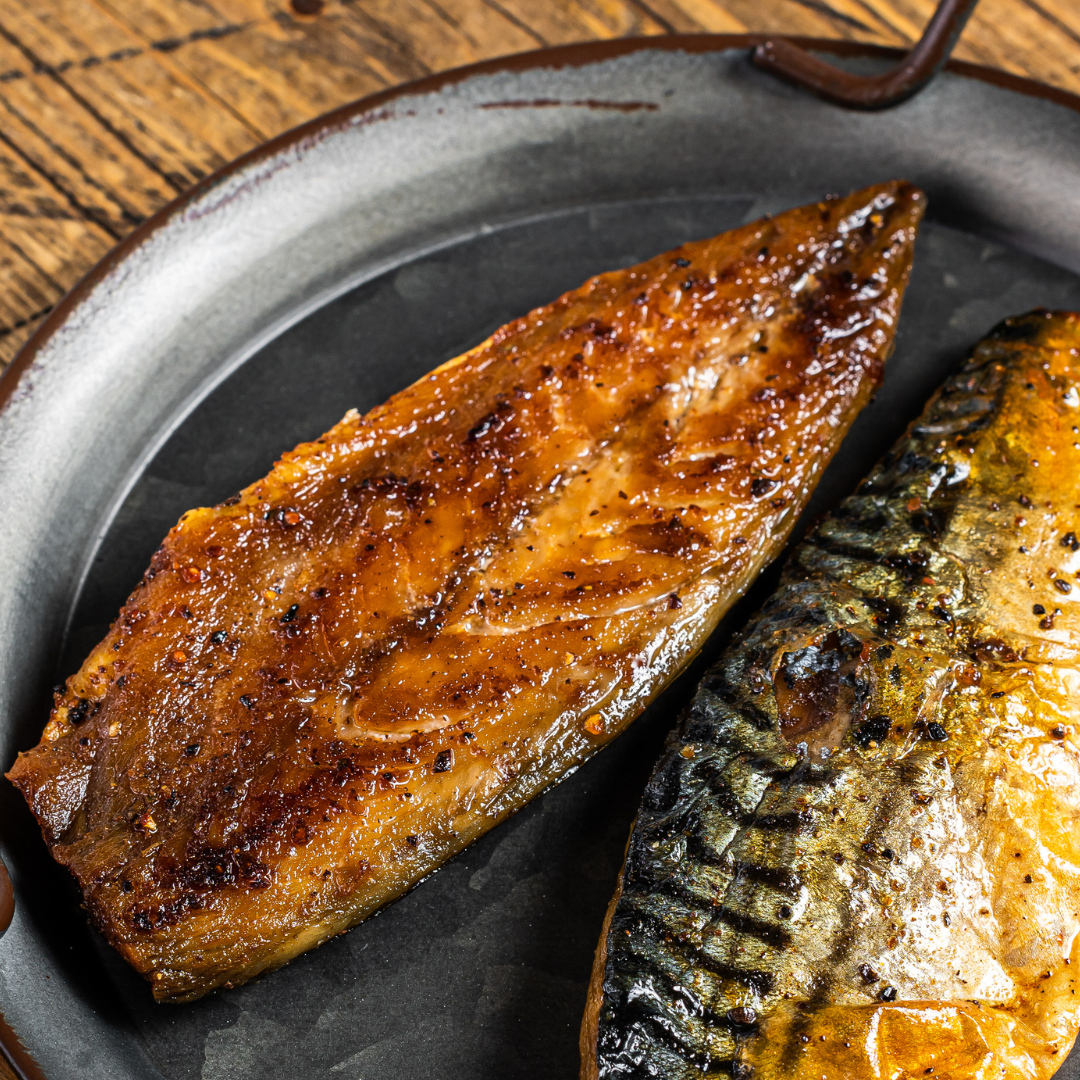 Two pieces of barbecue grilled saba mackerel fish fillet on a cast iron grill pan on a wooden surface table. 