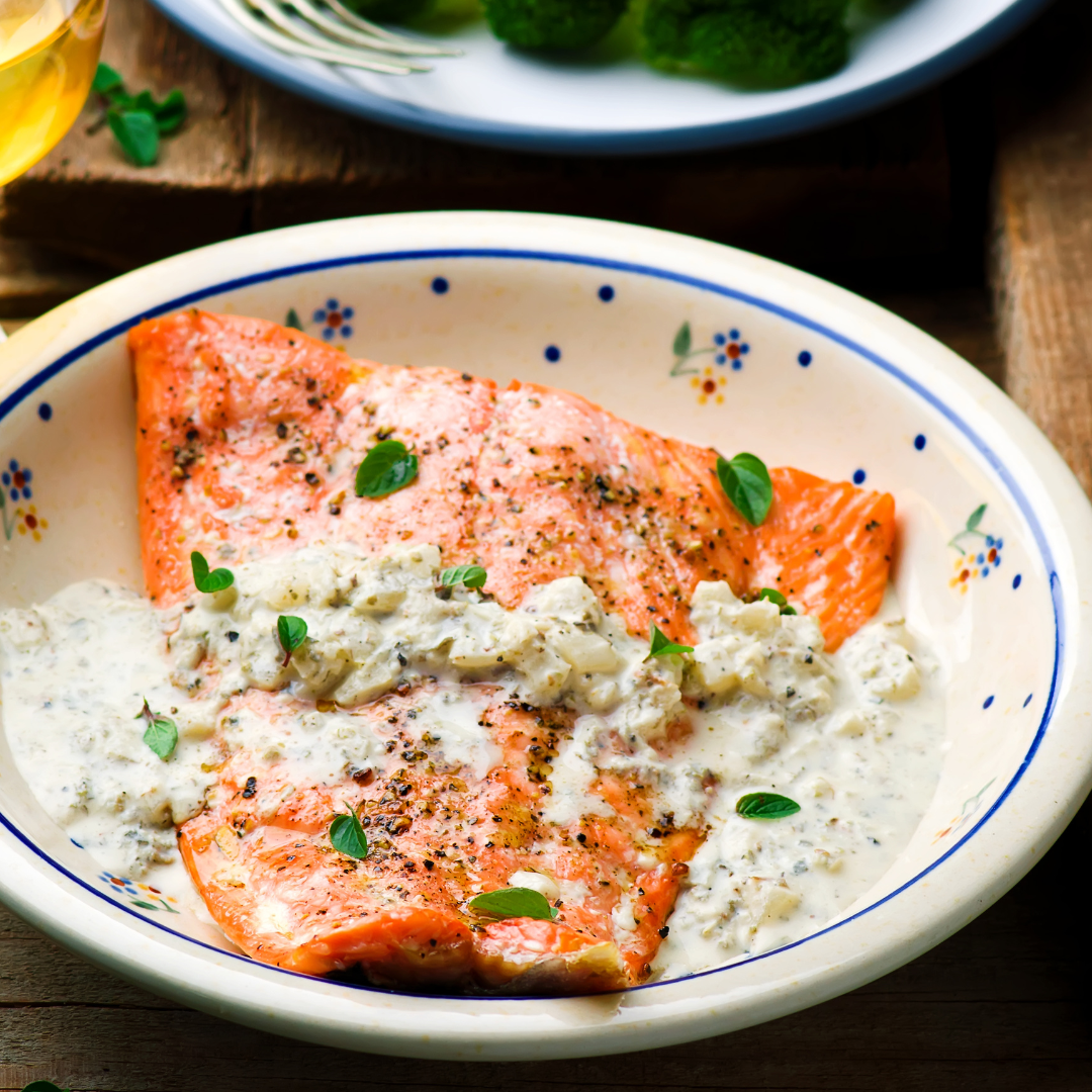 Seared salmon fillet topped with creamy garlic herb sauce, garnished with oregano on a ceramic plate.