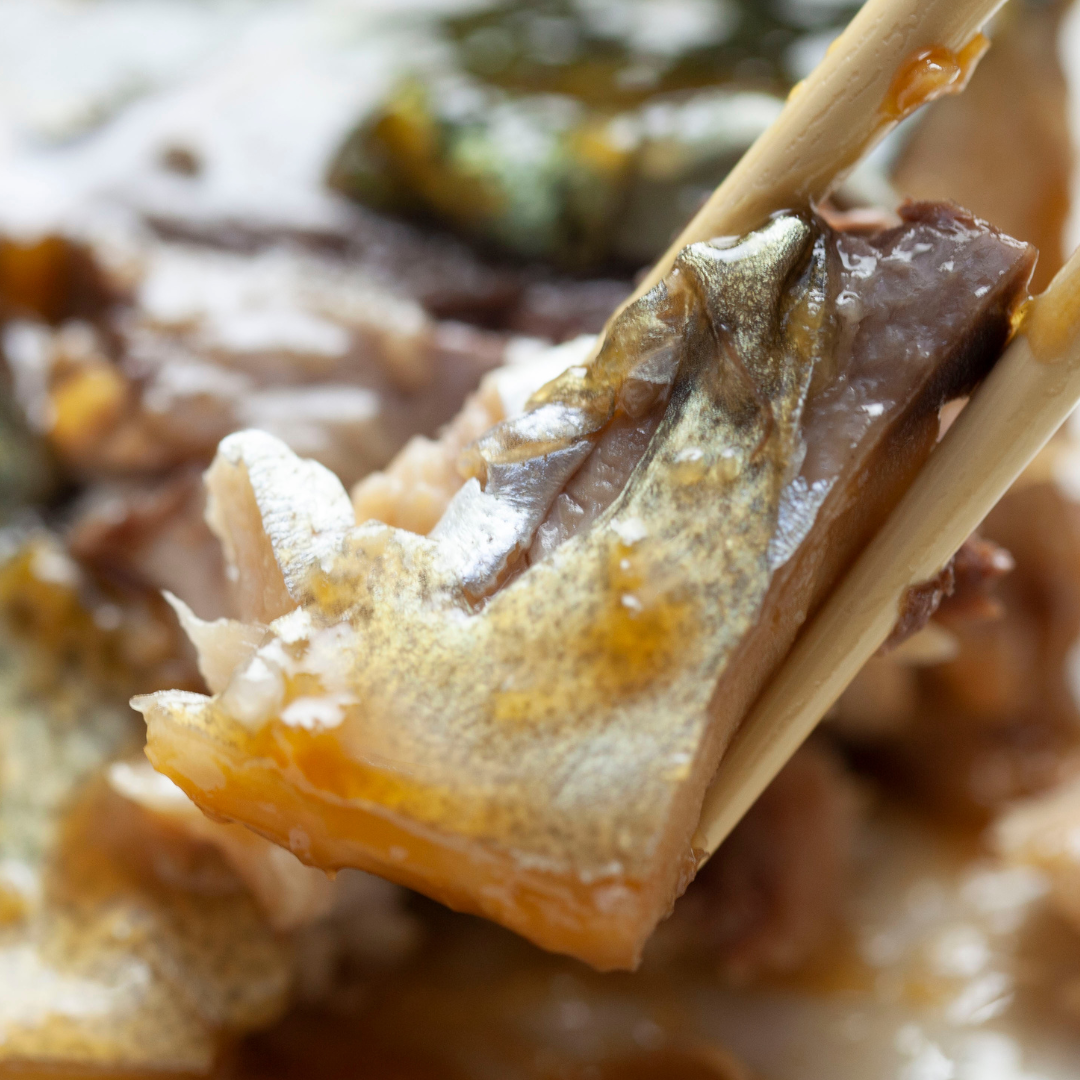 Close-up of a piece of miso grill saba mackerel fish meat (saba misoni) held with chopsticks over a dish with broth and vegetables.