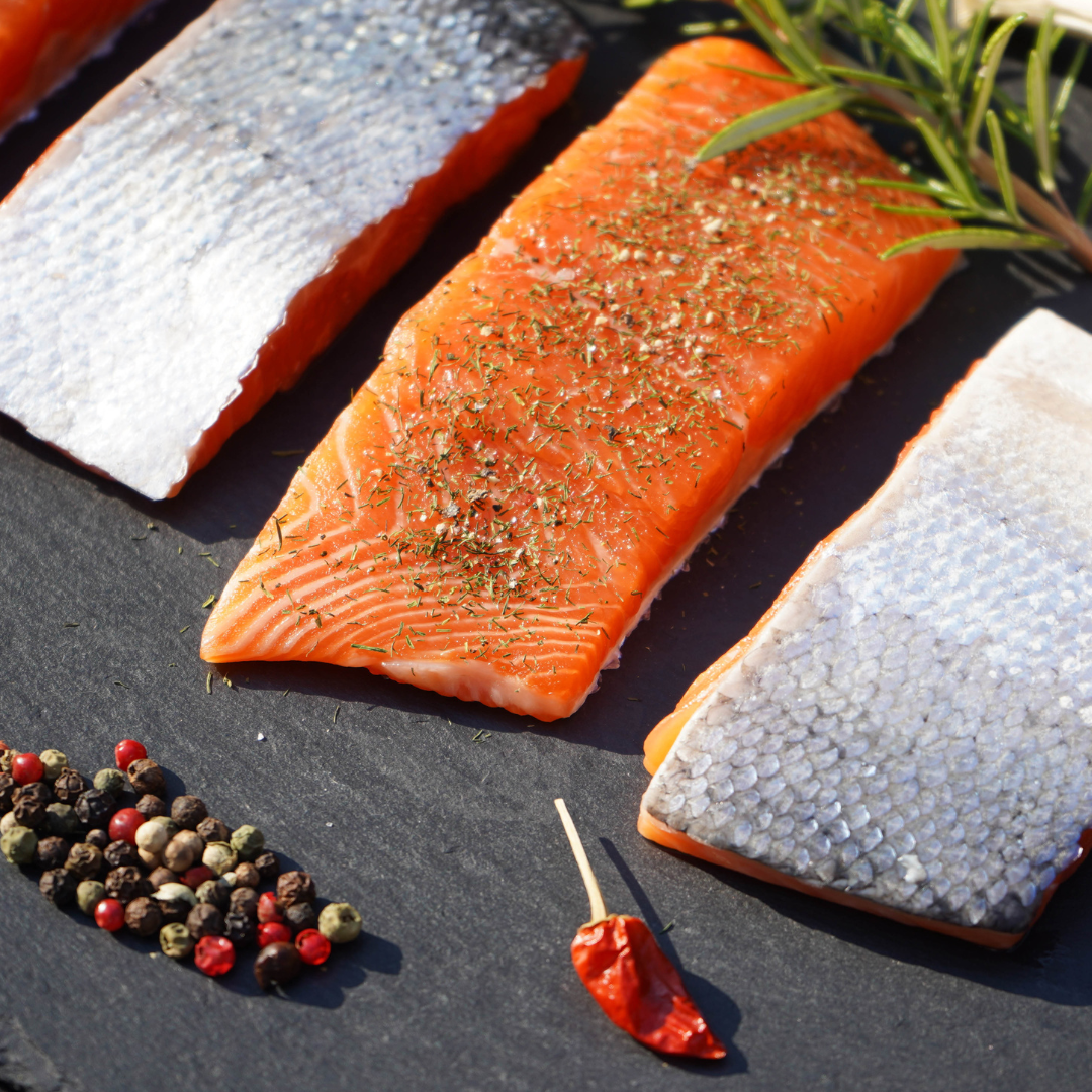 Skin-on salmon trout fillets seasoned with herbs and spices, displayed with peppercorns and chili on dark stone board